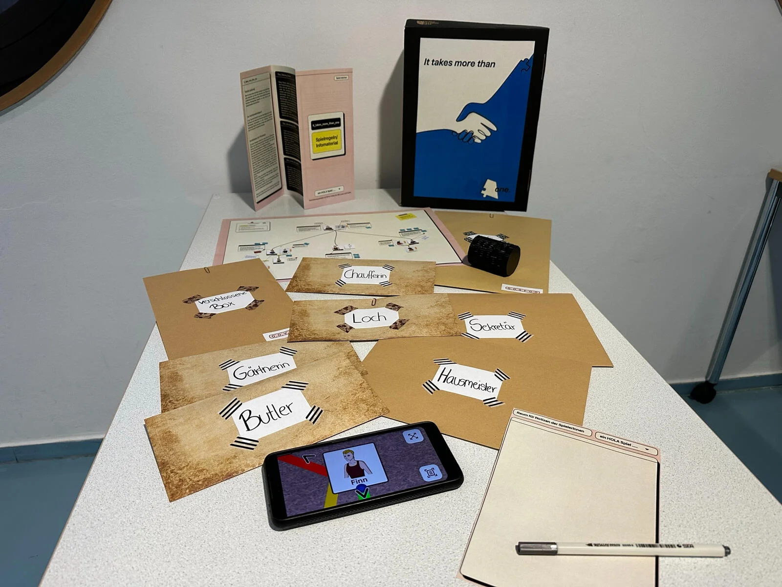 Complete HOLA game set laid out showing all physical components: character envelopes, clue documents, cipher wheel, and printed materials